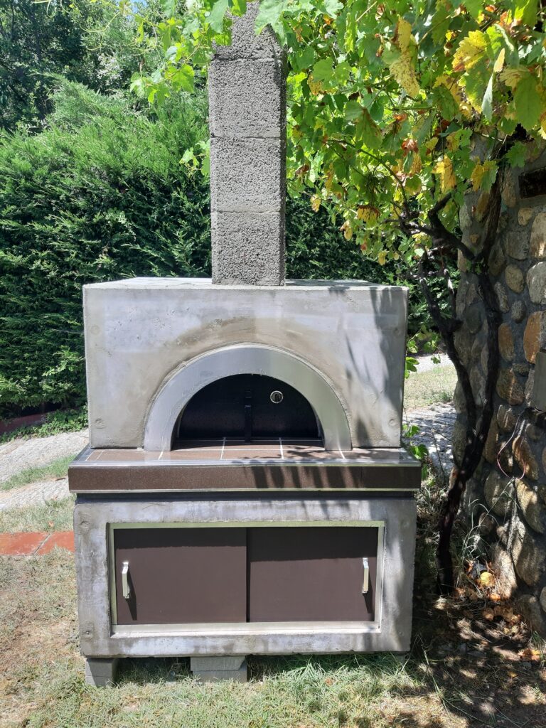 Traditional outdoor wood-fired pizza oven with a chimney, tiled countertop, and built-in storage cabinets.