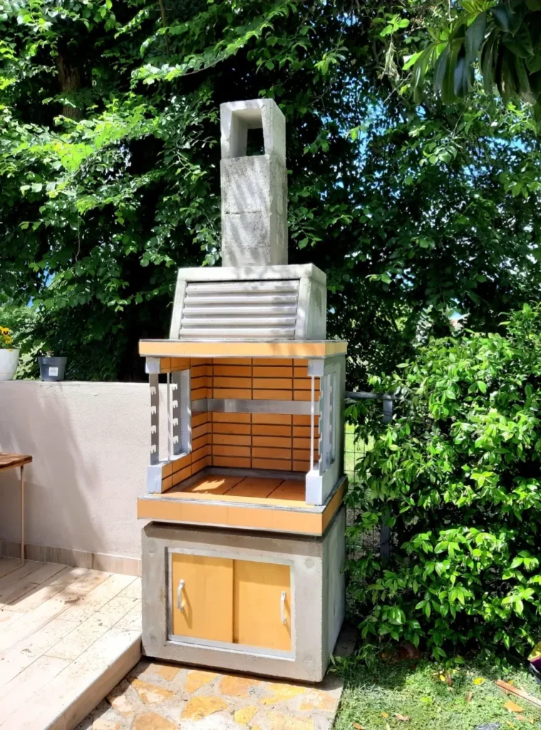 Compact outdoor barbecue with a chimney, brick-lined interior, and built-in storage cabinet, surrounded by lush greenery.