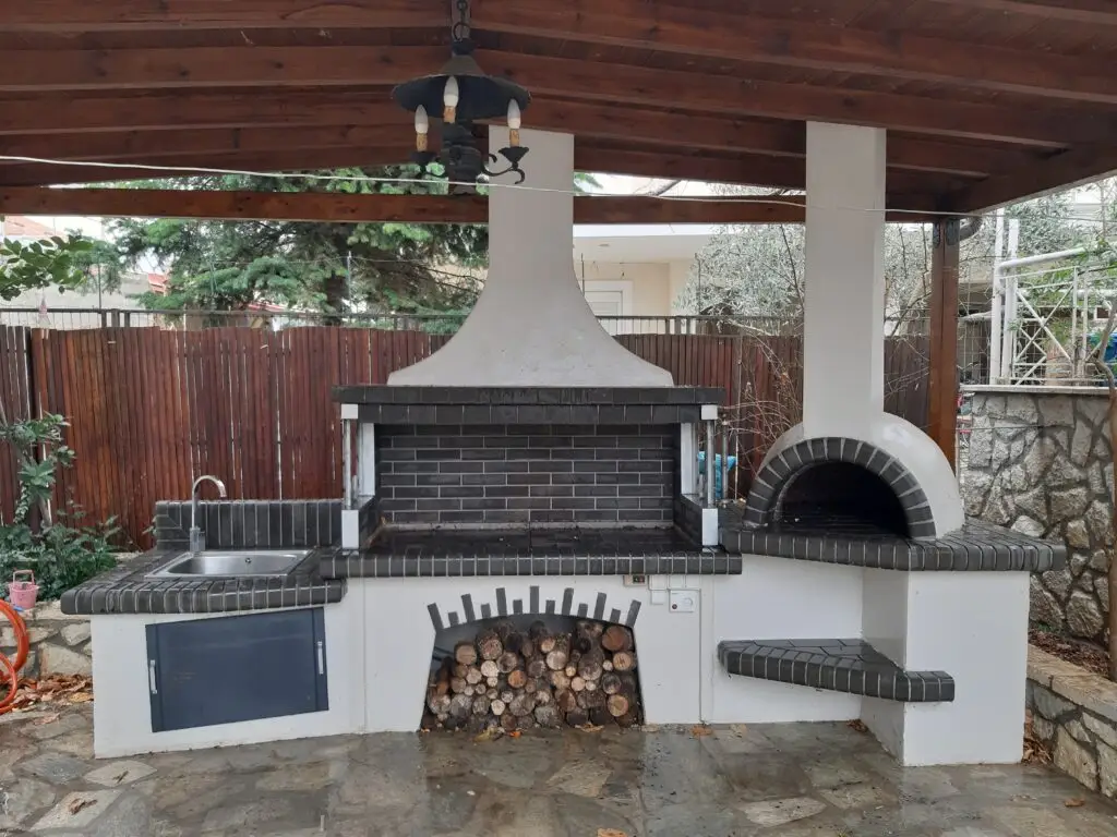 Rustic outdoor kitchen featuring a wood-fired pizza oven, barbecue grill, and integrated sink under a wooden pergola.